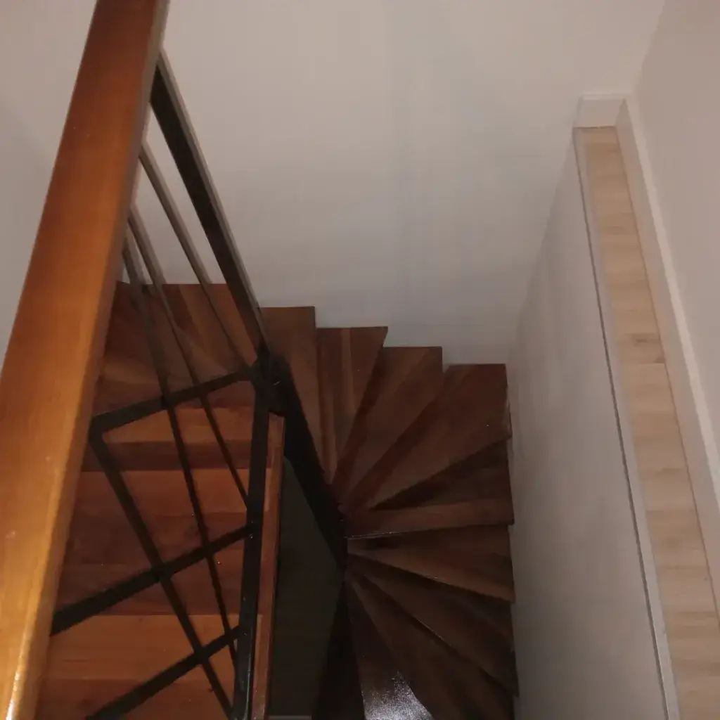 Kružne unutrašnje stepenice sa gazištem od punog drveta tople crvenkaste boje sa crnom metalnom ogradom i drvenim rukohvatom.
